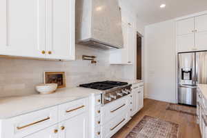 Kitchen with white cabinetry, stainless steel appliances, wall chimney range hood, light stone counters, and light wood-type flooring