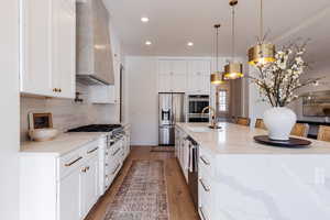 Kitchen featuring wall chimney range hood, hanging light fixtures, light stone counters, white cabinetry, and stainless steel appliances