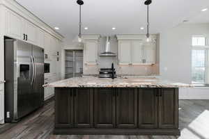 Kitchen with a center island with sink, black range with electric stovetop, wall chimney range hood, decorative light fixtures, and stainless steel fridge with ice dispenser