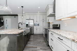 Kitchen featuring light stone counters, stainless steel appliances, wall chimney range hood, pendant lighting, and a center island with sink