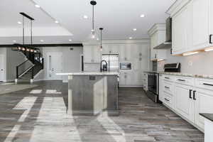 Kitchen featuring light stone counters, wall chimney exhaust hood, stainless steel appliances, decorative light fixtures, and an island with sink