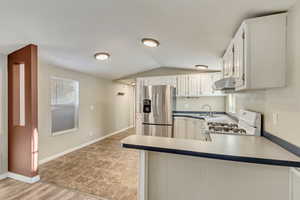 Kitchen featuring white gas stove, stainless steel fridge with ice dispenser, kitchen peninsula, lofted ceiling, and white cabinets