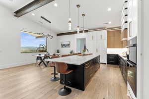 Kitchen with light stone counters, an island with sink, decorative light fixtures, white cabinets, and light wood-type flooring