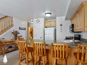 Kitchen with carpet flooring, a breakfast bar, kitchen peninsula, white fridge, and light brown cabinetry