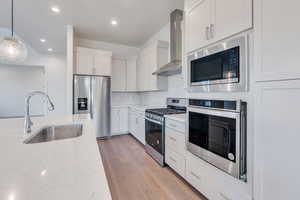 Kitchen featuring appliances with stainless steel finishes, sink, wall chimney range hood, white cabinetry, and hanging light fixtures