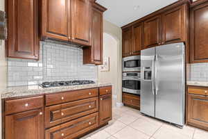 Kitchen with light stone countertops, range hood, decorative backsplash, light tile patterned floors, and appliances with stainless steel finishes