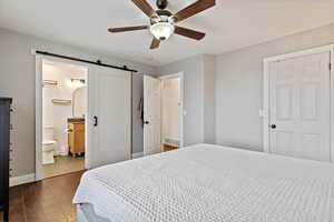 Bedroom with a barn door, ceiling fan, ensuite bath, and dark wood-type flooring