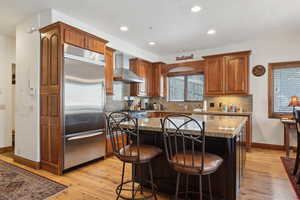 Kitchen featuring stainless steel appliances, wall chimney range hood, stone counters, a center island, and light hardwood / wood-style floors