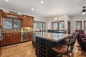 Kitchen with light stone countertops, light wood-type flooring, stainless steel dishwasher, decorative light fixtures, and a kitchen island