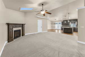 Unfurnished living room with a fireplace, light carpet, ceiling fan, and vaulted ceiling with skylight