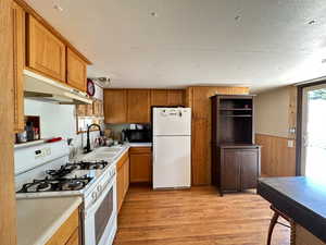 Kitchen with a textured ceiling, white appliances, wooden walls, sink, and light hardwood / wood-style flooring