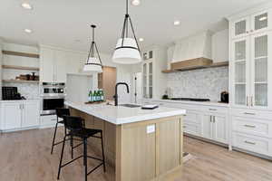 Kitchen featuring decorative backsplash, an island with sink, double oven, decorative light fixtures, and custom range hood