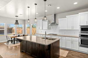 Kitchen featuring light stone countertops, stainless steel appliances, wall chimney range hood, a kitchen island with sink, and white cabinets