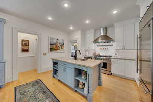 Kitchen with white cabinetry, high end appliances, wall chimney range hood, and an island with sink