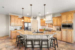 Kitchen featuring a breakfast bar, custom range hood, stainless steel appliances, decorative light fixtures, and a center island with sink