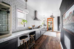 Kitchen with light stone counters, dark wood-type flooring, pendant lighting, wall chimney range hood, and stainless steel gas cooktop