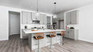 Kitchen with appliances with stainless steel finishes, gray cabinetry, a kitchen island with sink, wood-type flooring, and decorative light fixtures