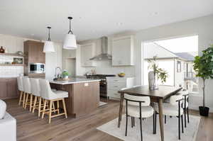 Kitchen featuring an island with sink, stainless steel appliances, wall chimney range hood, decorative backsplash, and white cabinetry