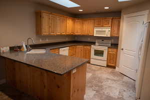 Kitchen featuring sink, white appliances, dark stone countertops, kitchen peninsula, and a skylight