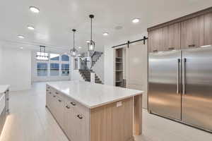 Kitchen with a barn door, hanging light fixtures, stainless steel built in fridge, light stone counters, and a kitchen island