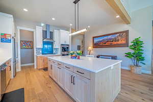 Kitchen featuring wall chimney exhaust hood, a spacious island, decorative light fixtures, decorative backsplash, and light brown cabinetry