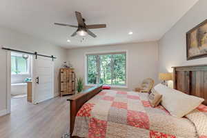 Bedroom with ceiling fan, ensuite bath, light wood-type flooring, and a barn door