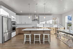 Kitchen with appliances with stainless steel finishes, wall chimney exhaust hood, a kitchen island with sink, white cabinets, and hanging light fixtures