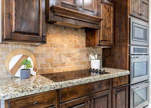 Kitchen with light stone counters, stainless steel appliances, custom range hood, backsplash, and dark brown cabinetry