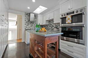 Kitchen with white cabinets, double oven, wall chimney exhaust hood, a skylight, and dark hardwood / wood-style flooring