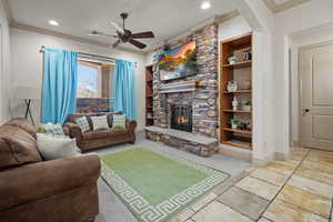 Living room with ornamental molding, ceiling fan, built in features, and a stone fireplace