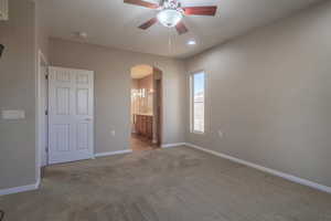 Unfurnished primary bedroom featuring connected bathroom, ceiling fan, and light colored carpet