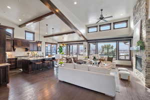 Living room with vaulted ceiling with beams, ceiling fan with notable chandelier, a stone fireplace, and dark wood-type flooring