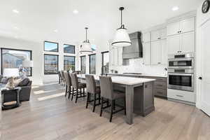 Kitchen featuring custom exhaust hood, an island with sink, appliances with stainless steel finishes, light wood-type flooring, and pendant lighting