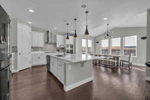 Kitchen featuring lofted ceiling, white cabinetry, wall chimney range hood, sink, and a center island with sink