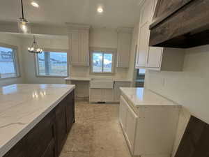 Kitchen featuring range hood, hanging light fixtures, white cabinets, light stone counters, and a chandelier