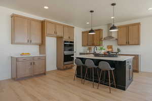Kitchen with wall chimney range hood, stainless steel double oven, a kitchen island with sink, and light wood-type flooring