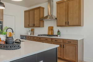 Kitchen with stainless steel gas cooktop, brown cabinets, wall chimney range hood, and light countertops