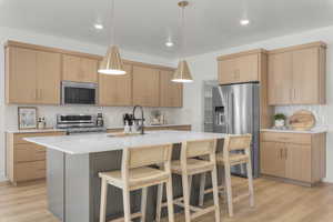 Kitchen featuring an island with sink, stainless steel appliances, decorative backsplash, decorative light fixtures, and light brown cabinetry