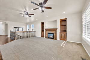 Unfurnished living room with ceiling fan, built in shelves, dark colored carpet, and a fireplace