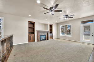 Unfurnished living room with ceiling fan, built in shelves, a tile fireplace, and light colored carpet