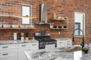 Kitchen with brick wall, wall chimney exhaust hood, sink, light stone counters, and stainless steel range with gas stovetop