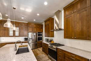 Kitchen featuring appliances with stainless steel finishes, pendant lighting, wall chimney exhaust hood, light stone counters, and sink