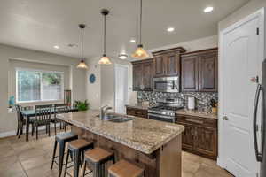 Kitchen featuring a center island with sink, appliances with stainless steel finishes, decorative light fixtures, light stone counters, and sink