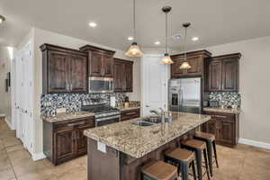 Kitchen featuring decorative light fixtures, stainless steel appliances, an island with sink, sink, and light stone counters