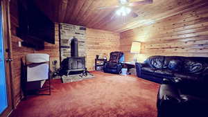 Living room with carpet, ceiling fan, wooden walls, a wood stove, and wooden ceiling