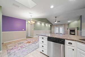 Kitchen with white cabinetry, vaulted ceiling with skylight, dishwasher, light stone countertops, and ceiling fan with notable chandelier