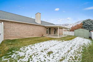 Snow covered back of property with a lawn, a mountain view, a storage unit, and a patio