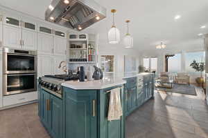 Kitchen featuring appliances with stainless steel finishes, white cabinetry, wall chimney range hood, hanging light fixtures, and a kitchen island with sink