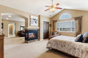 Primary suite bedroom featuring ceiling fan, light colored carpet, a multi sided fireplace, and vaulted ceiling