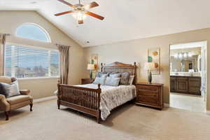 Primary suite bedroom featuring ceiling fan, light colored carpet, connected bathroom, and vaulted ceiling